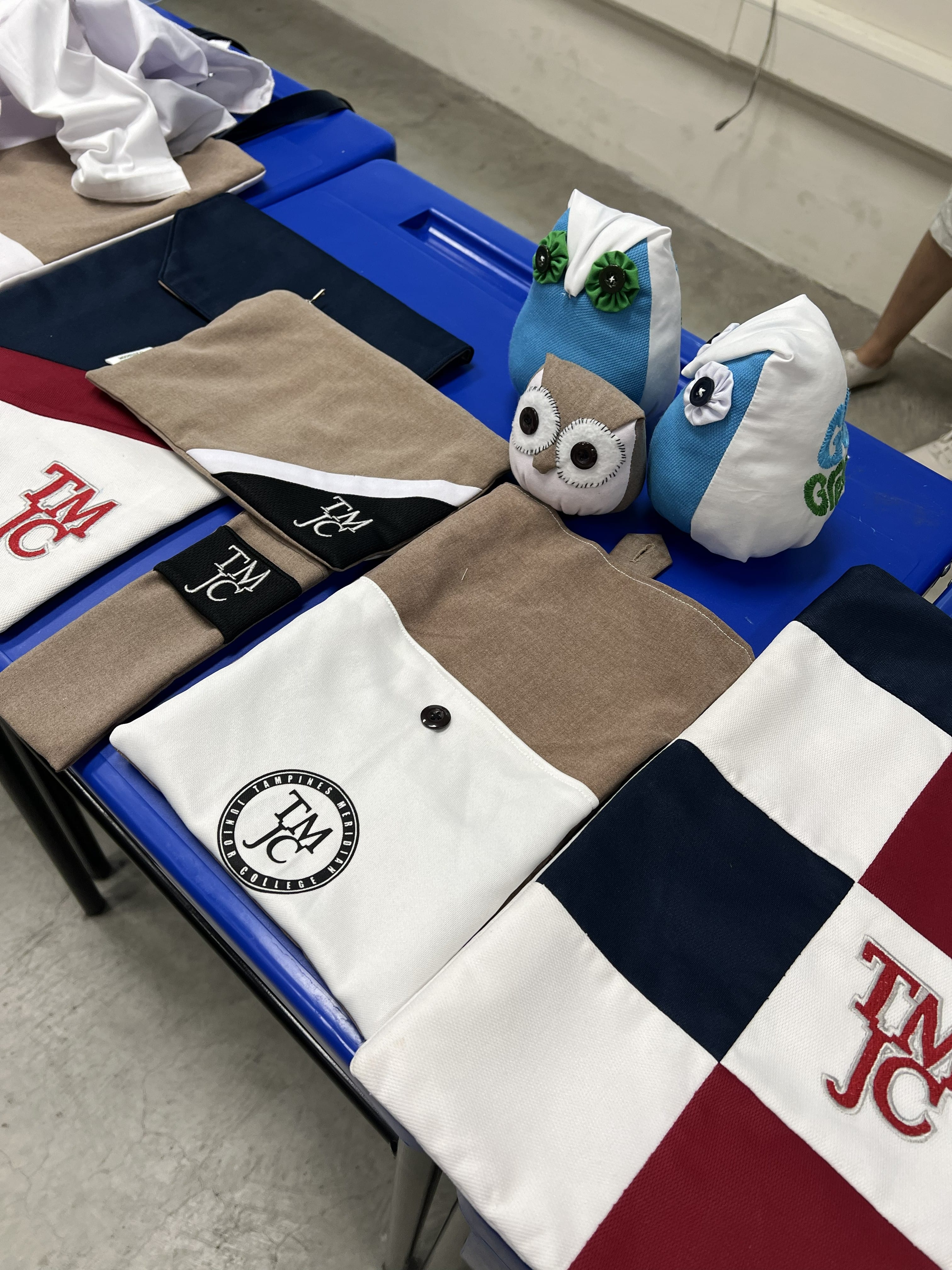 A table displays handmade fabric items, including pouches and tote bags decorated with the "TMJC" school logo, alongside two small stuffed owl toys.